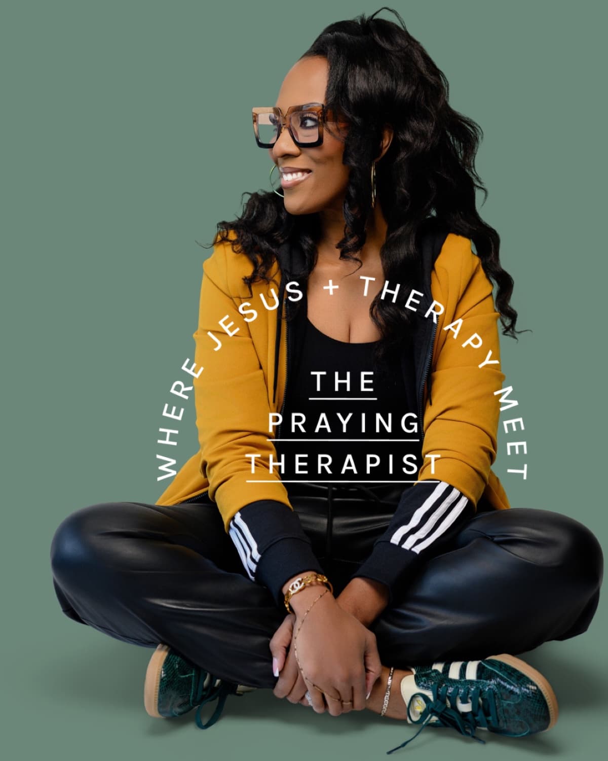 Yvonne Sailor-Walker seated cross-legged against a sage green backdrop in a mustard jacket
